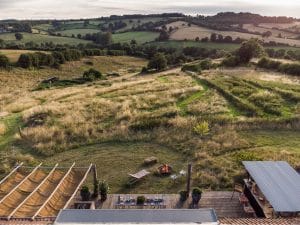Aerial view of a grassy hillside garden with a wooden deck, outdoor seating, a small fire pit, and rolling fields in the background—your perfect summer oasis for relaxing in all moments.