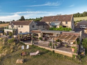 A large stone house with a spacious wooden deck, outdoor seating, and garden offers the perfect Summer Oasis, set in a rural landscape with fields and trees under a blue sky.