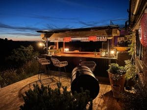 An outdoor bar with stools, hanging glasses, and warm lighting sits on a wooden deck at dusk, creating a Summer Oasis surrounded by plants and overlooking a dark landscape.