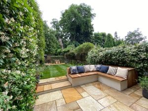 A contemporary landscaped backyard with a paved patio, L-shaped wooden bench with cushions, and a lawn bordered by hedges and trees.