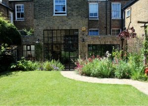A chillout garden with a curved stone path leads to a brick house featuring large black-framed glass doors and windows. Flowerbeds and greenery border the lush lawn.