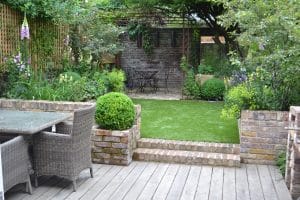 A neatly landscaped backyard with a wooden deck, floating bench, wicker dining set, brick planters, artificial lawn, and a small seating area under a pergola surrounded by greenery.