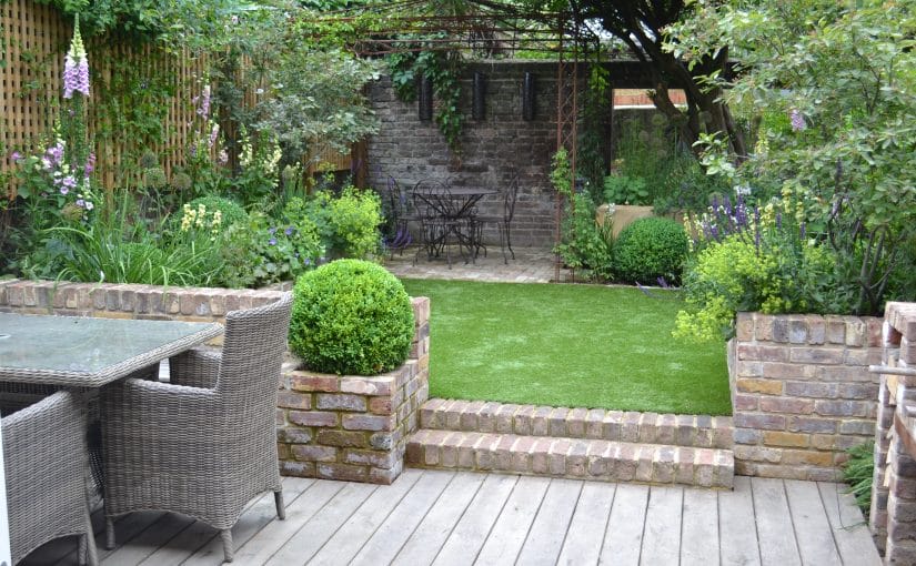 A neatly landscaped backyard with a wooden deck, floating bench, wicker dining set, brick planters, artificial lawn, and a small seating area under a pergola surrounded by greenery.