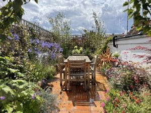 Wooden table and chairs are set on a tiled patio in the Portobello Sky Garden, surrounded by lush plants and flowers under a partly cloudy sky.