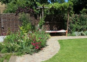 A chillout garden featuring a curved stone path, lush lawn, vibrant flower beds, and a wooden arbour with a bench beneath woven fencing in the background.