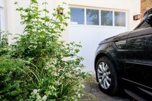 A black SUV is parked in front of a white garage door, beside green plants with white flowers in the front garden.