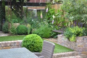 A landscaped garden with green shrubs, flowering plants, a brick retaining wall, a floating bench, a wicker chair, and a glass-topped table in the foreground.