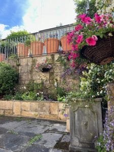 Stone wall with flower pots on top, hanging basket and climbing plants buzzing with bees, and a paved area with greenery under a partly cloudy sky.