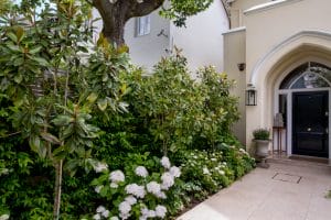 A paved walkway winds through the front garden, leading to a black front door with an arched entryway, surrounded by lush green shrubs and blooming white flowers.