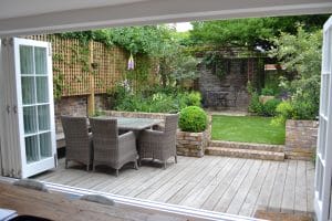 View from an open doorway showing a patio with a wicker dining set, floating bench, wooden deck, steps, and a landscaped garden with greenery and brick walls.