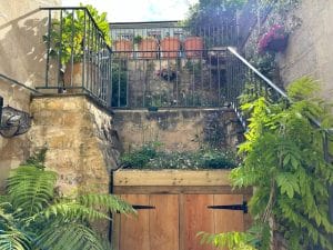 Two wooden doors are set into a stone wall below a garden terrace buzzing with bees, potted plants, metal railings, and greenery on both levels. Sunlight illuminates the lively scene.