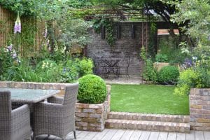 A small, neatly landscaped garden with a patio seating area, artificial grass, and a floating bench alongside metal table and chairs near a brick wall, all surrounded by lush greenery and flowers.