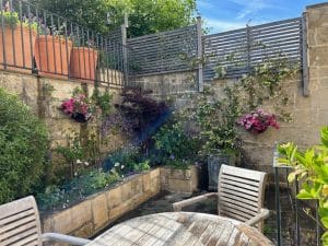Small patio garden with wooden chairs and table, potted plants on a ledge, climbing vines, and hanging flower baskets attracting bees against a stone wall under a blue sky.