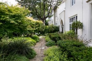 A stone pathway winds through a lush front garden beside a white residential building with multiple windows and elegant arched doorways.