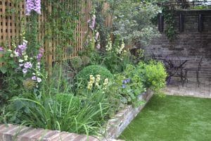 A garden featuring a brick-edged flower bed, lush green plants, purple flowers, a grassy lawn, and a modern floating bench alongside a metal table with chairs beside the brick wall.