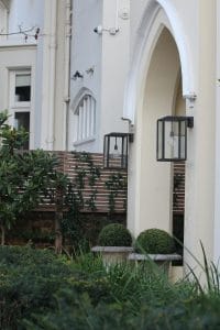 Cream-colored building exterior with arched doorway, modern black lantern, potted spherical shrubs, wooden fence, and lush front garden foliage in the foreground.