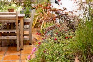 Wooden patio table and chairs sit on a tiled terrace in the Portobello Sky Garden, nestled beside a garden bed filled with lush green plants and vibrant red flowers.