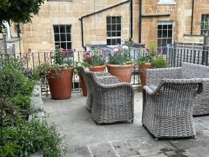 Four wicker chairs and a table are arranged on a stone patio with large potted plants and flowers, attracting gentle bees, set against a beige brick building with windows.