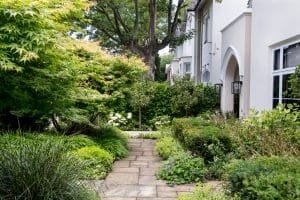A stone pathway winds through a lush front garden, guiding you to the entrance of a white house with arched doorway and windows.