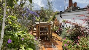 A wooden table and chairs sit on a wet patio surrounded by lush plants and flowers, reminiscent of a Portobello Sky Garden, with a white wall and chimneys in the background under a partly cloudy sky.