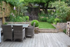 A wooden deck with a wicker dining set and a sleek floating bench overlooks a small lawn bordered by brick walls and lush, green garden beds.
