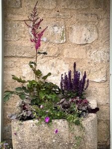 A stone planter against a brick wall contains various flowering plants, including tall pink and purple blooms with green foliage that attract bees.