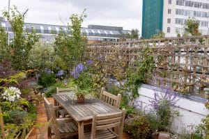A wooden table with chairs is set on the Portobello Sky Garden rooftop, surrounded by lush plants and flowers, with city buildings in the background.