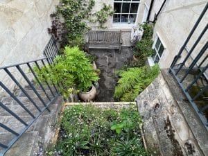 A small stone courtyard with a wooden bench, potted plants, ferns, and greenery attracts bees, all enclosed by stone walls and metal railings.