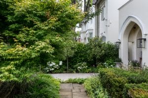 A stone pathway winds through a beautifully landscaped front garden filled with green shrubs and trees, set before charming white, two-story houses.
