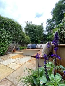 Symmetrical stone patio with cushioned seating, striped pillows, and potted purple flowers in the foreground, surrounded by green hedges and trees under a cloudy sky.