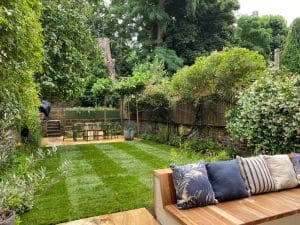 A well-maintained, symmetrical garden with a lush lawn, outdoor dining table and chairs, vibrant greenery, and a wooden bench with patterned cushions in the foreground.