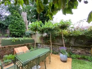 A symmetrical outdoor garden with a green metal dining table and chairs, potted plants, a striped lounge chair, and leafy trees arranged by a brick wall and wooden fence.