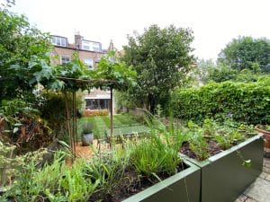 A landscaped, symmetrical garden with green hedges, a lawn, raised planters filled with various plants, and a multi-story brick building in the background.