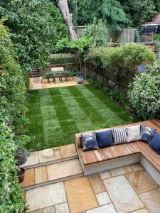 A landscaped garden with a symmetrical checkered lawn, patio seating with cushions, potted plants, and an outdoor dining table in the background.