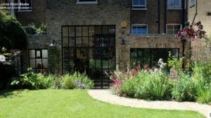 A modern brick house with large black-framed glass doors, this Exotic Retreat is surrounded by a lush garden of colorful flowering plants and a curved stone path, basking in the sunshine.