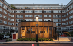 Contemporary single-story building with expansive windows and warm lighting stands before a multi-story brick apartment complex, framed by a streetlamp and parked cars.