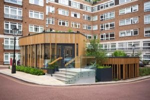 A contemporary glass and wood extension with steps and a sleek glass railing is attached to a multi-story brick apartment building with many windows.