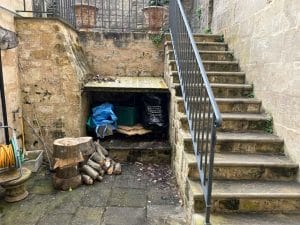 A stone courtyard with a stack of firewood and logs beside a covered storage area under outdoor stairs. A hose and potted plants are also visible, with bees occasionally buzzing among the greenery.
