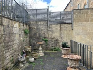 A small, enclosed stone patio with potted plants, a birdbath, a rooster statue, and some ivy growing on the walls attracts gentle bees buzzing among the blossoms.