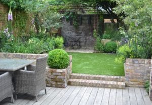 An inviting outdoor garden with a wooden deck, wicker dining set, manicured lawn, brick planters, and a cozy seating area under trees at the back.