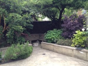 A small front garden patio with raised beds, various green and purple leafy plants, and a wooden bench set against a brick wall.