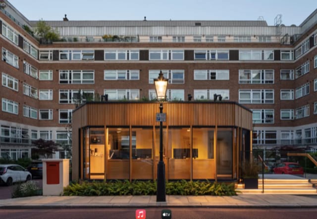 A contemporary glass and wood structure sits in front of a multi-story brick apartment building, illuminated at dusk with a lamppost and red mailbox nearby.