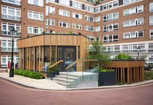 A contemporary building with glass walls and wooden slats is attached to a multi-story brick apartment complex, featuring a glass-railed staircase and landscaped plants around the entrance.