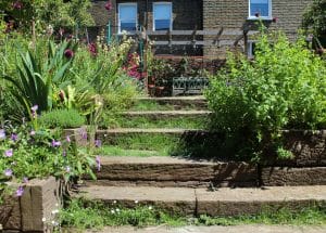 Stone steps wind through a lush chillout garden filled with green plants and flowers, guiding you toward a wooden pergola and a metal bench set before a charming brick building.
