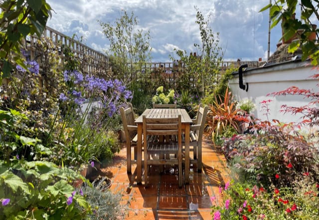 Wooden table and chairs are set on a tiled patio, surrounded by lush green plants and colorful flowers, under a partly cloudy sky.
