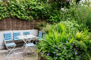 A small outdoor chillout garden seating area with a white table and metal chairs with blue cushions, surrounded by lush green plants and flowers.