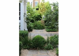 A symmetrical stone path leads through a lush, green garden with bushes, ornamental grasses, and a small tree beside a white building.