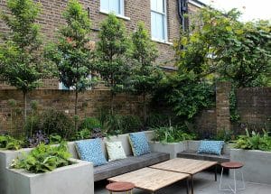 A modern, symmetrical garden patio featuring wooden benches with patterned cushions, a wooden coffee table, potted plants, and a brick wall backdrop adorned with greenery.