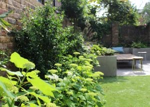 A landscaped backyard garden with symmetrical green plants along a brick wall, a concrete seating area, a wooden table, stools, and a cushioned bench.