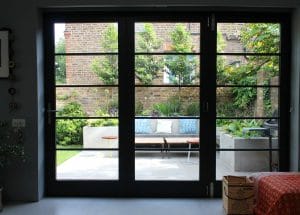 View through large glass doors reveals a symmetrical patio setup with a bench, stools, potted plants, and a brick wall in the background.